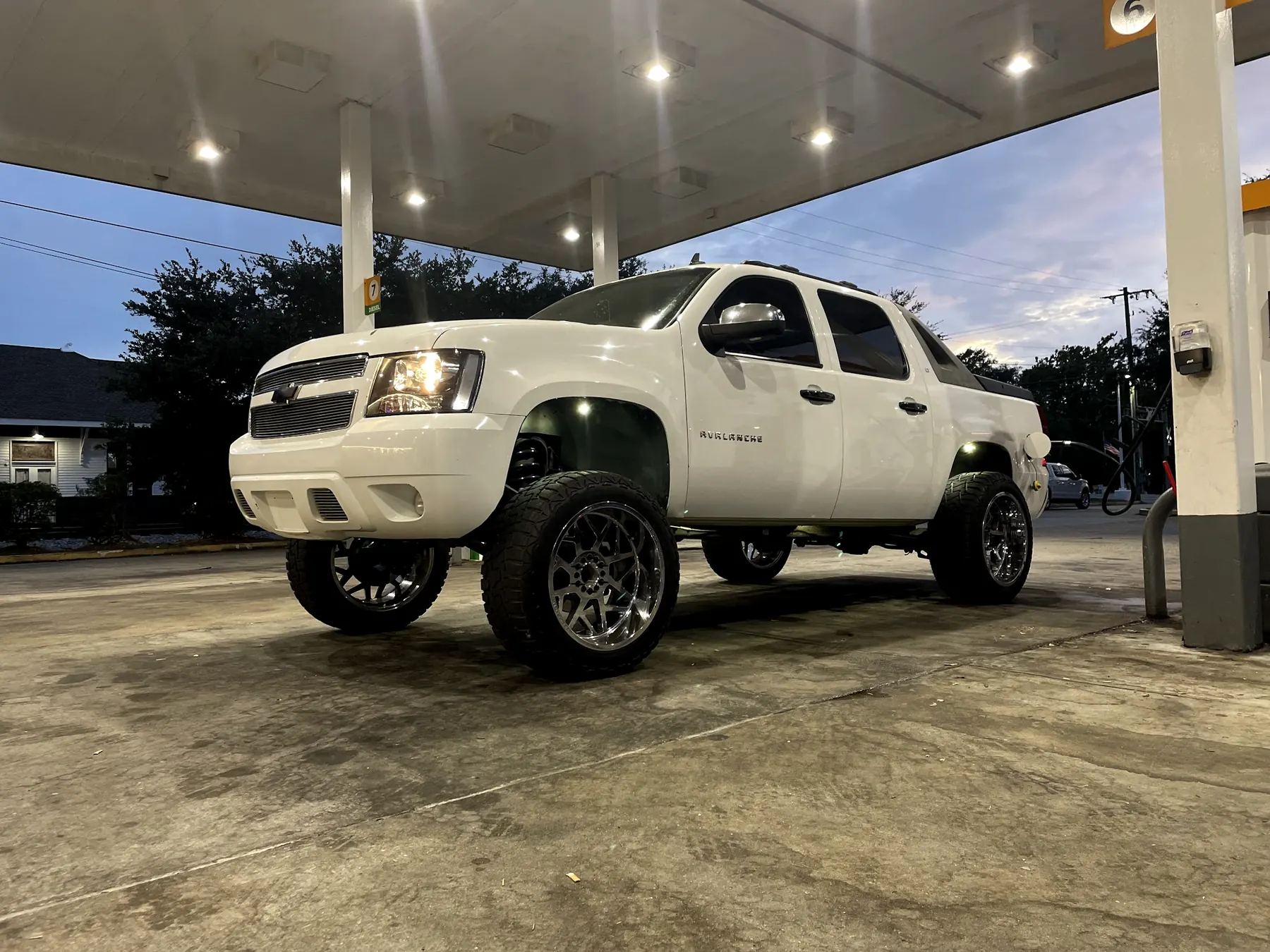 A white lifted Chevy Avalanche at a fuel canopy at dusk, its headlights on