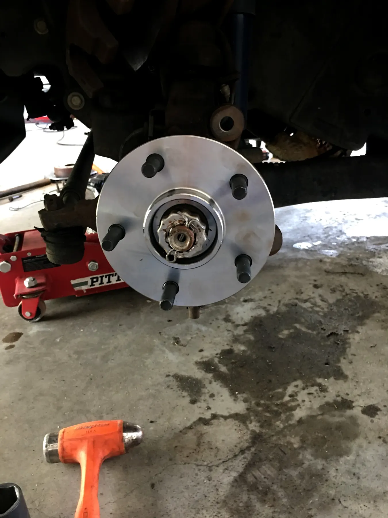 A wheel hub on a truck held up by a red floor jack, with an orange hammer on the shop floor
