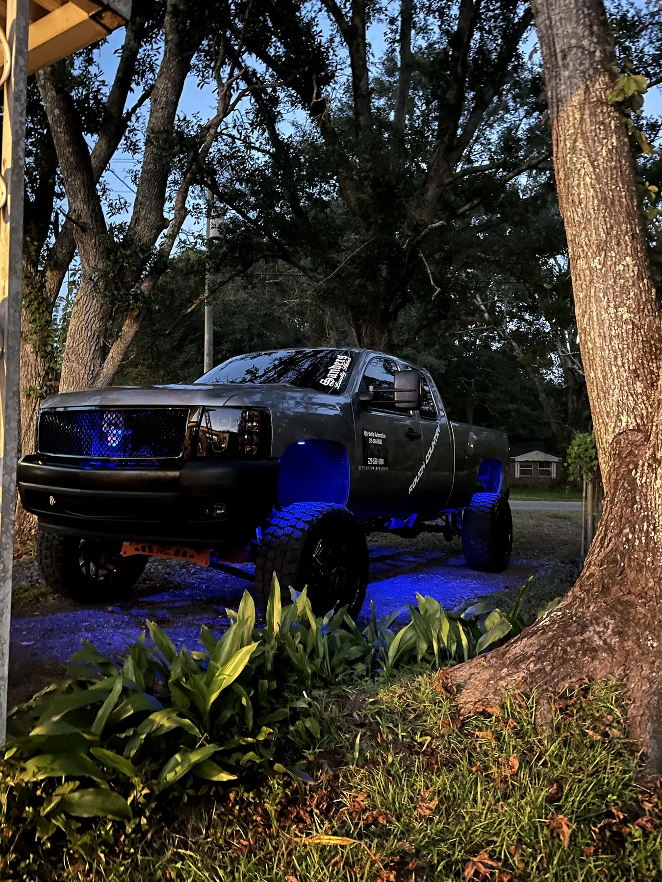 A lifted truck under oak trees at night, blue underglow lighting the ground