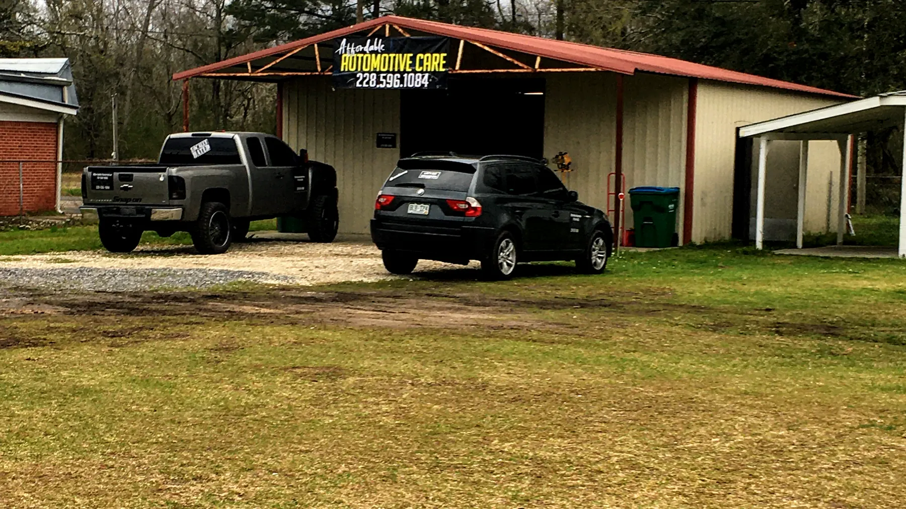 Two trucks parked in the grass in front of our shop under a big oak, the red roof and sign behind them